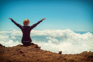 a woman sitting on the ground overlooking the clouds with her hands in the air in triumph. She has beaten alcohol abuse.