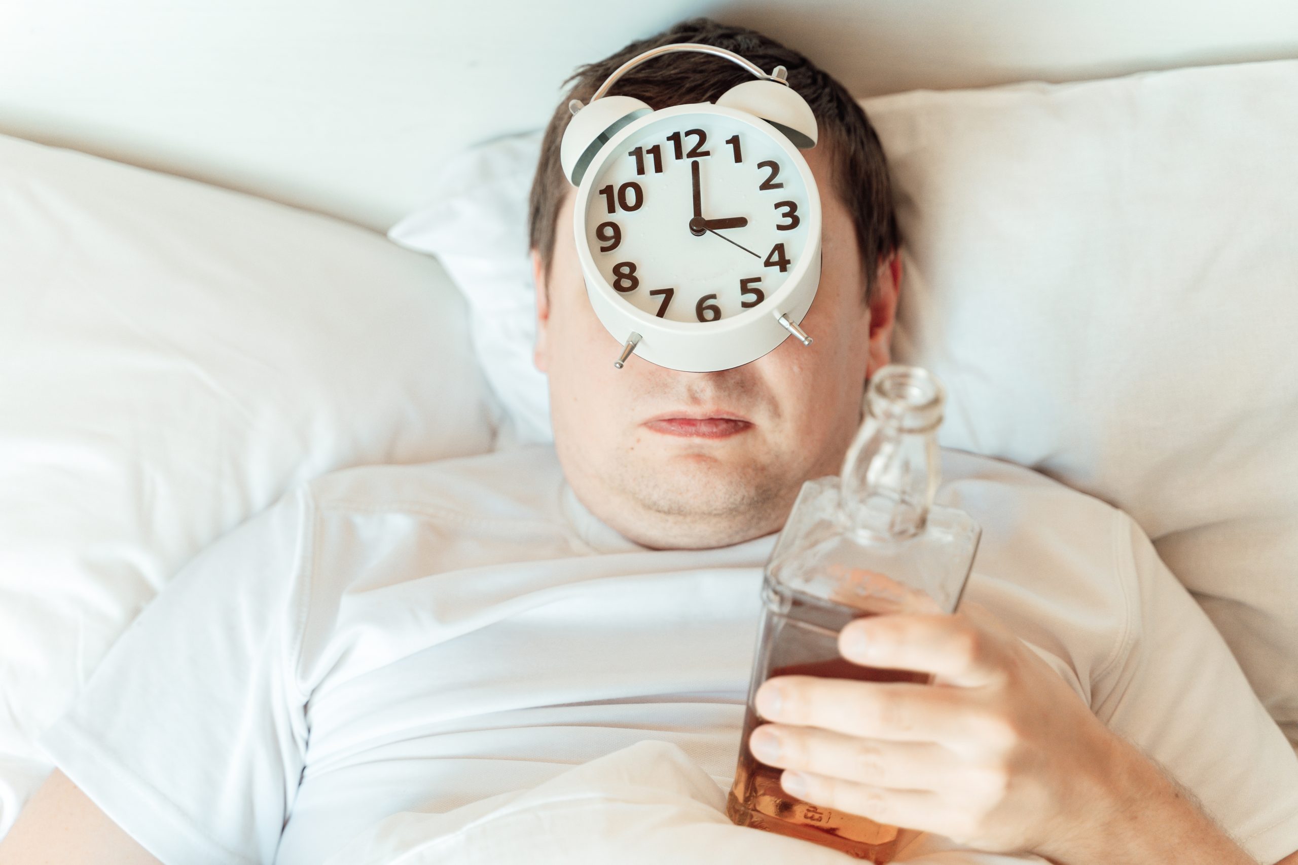 a man in bed holding a bottle of liquor with an alarm clock on his head. He's wondering how to sleep without alcohol