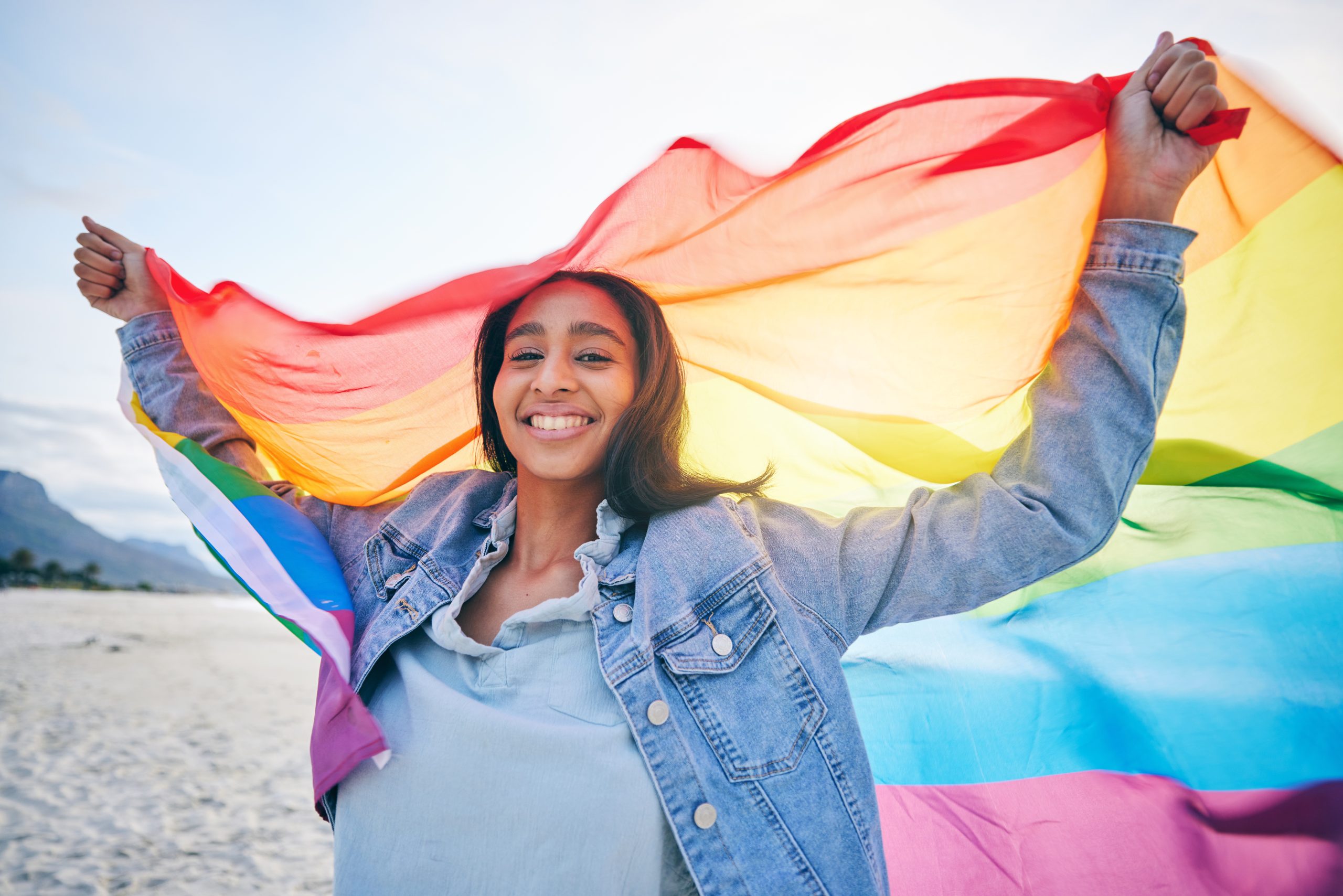 a woman on the beach holding a Pride flag behind her blowing in the wind to celebrate LGBTQ addiction treatment